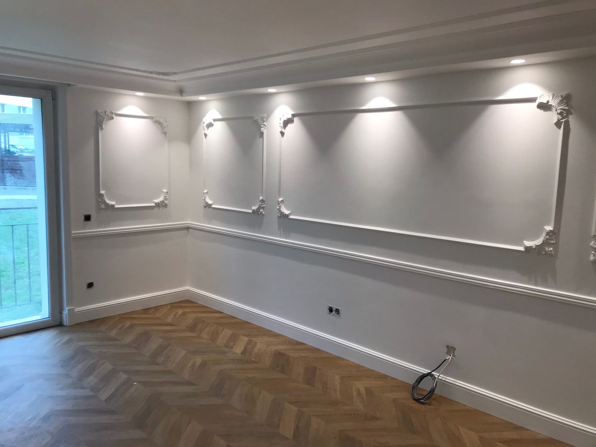 Jasne wnętrze po remoncie z dekoracyjnymi, białymi sztukateriami na ścianach, parkietem ułożonym w jodełkę, widoczną instalacją elektryczną i oknem balkonowym.