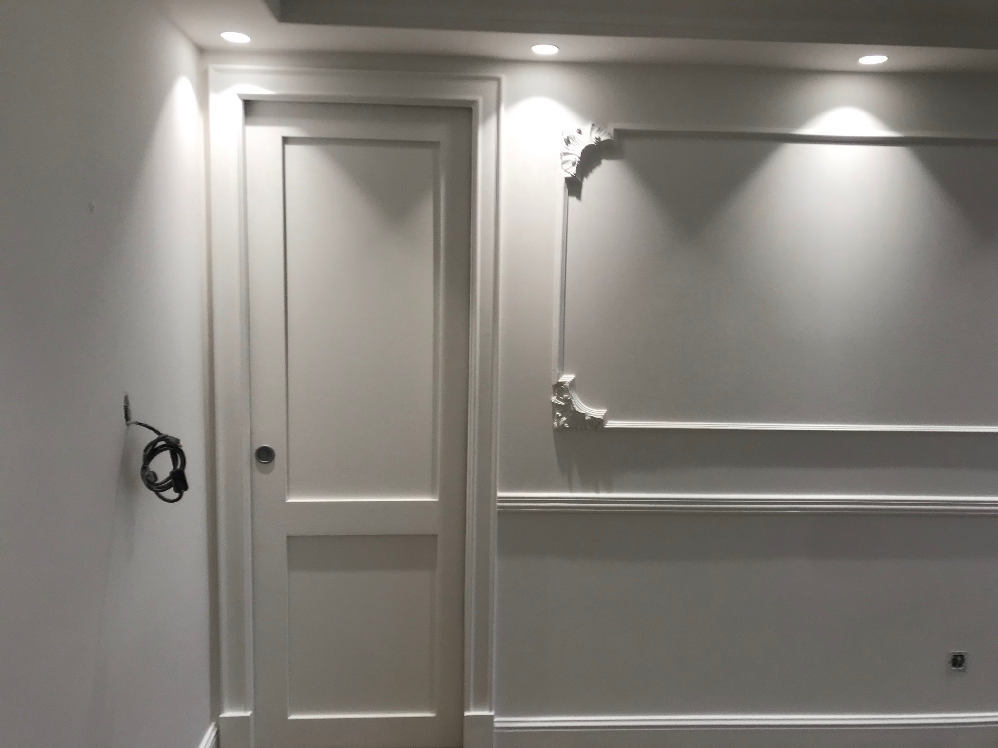 Białe, dwuskrzydłowe drzwi wewnętrzne z delikatnymi frezowaniami, umieszczone obok ściany z dekoracyjną sztukaterią w stylu klasycznym, oświetlone punktowymi lampami sufitowymi, z widocznymi...