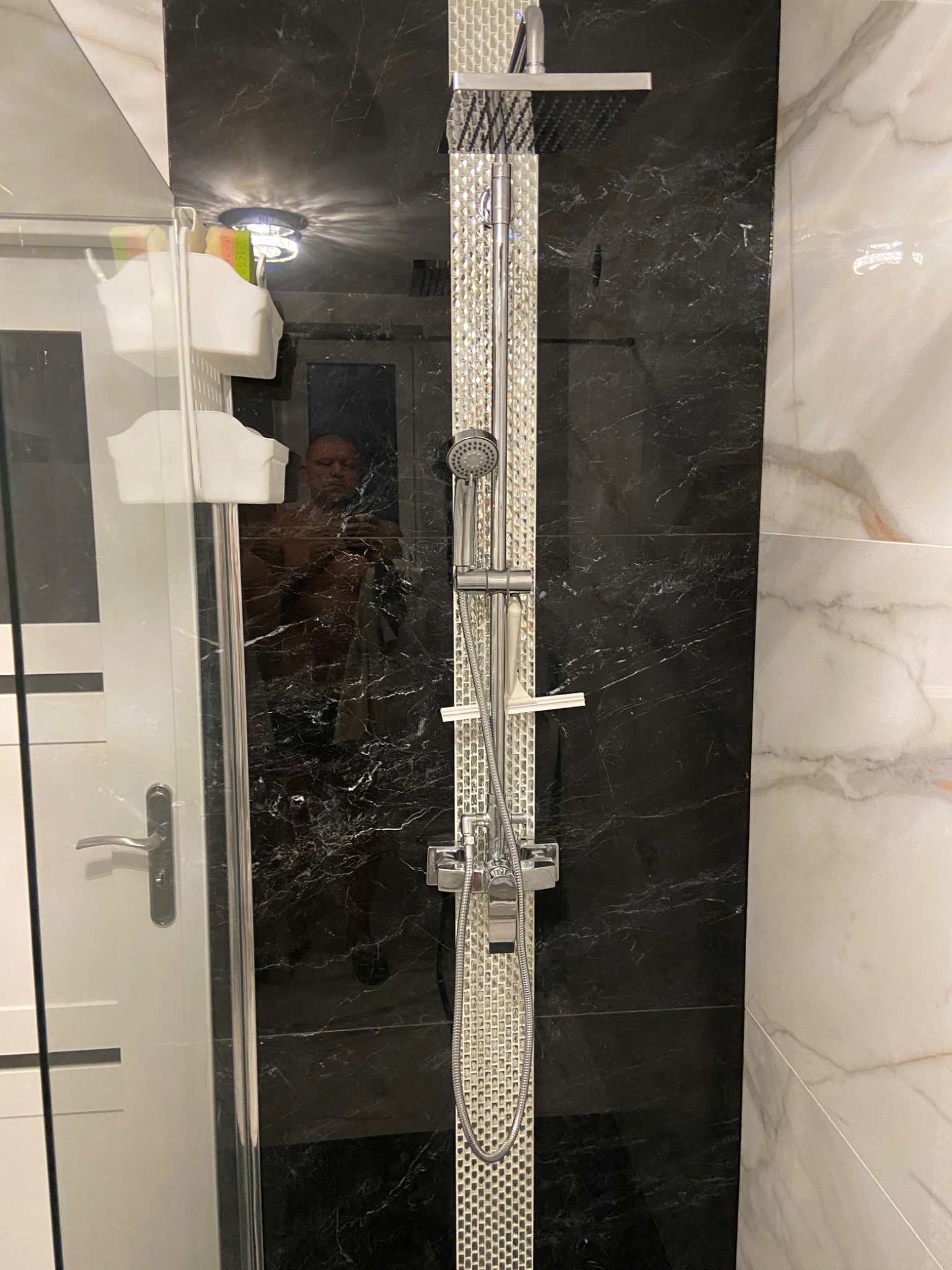 Nowoczesna kabina prysznicowa z czarną marmurową ścianą i srebrnym panelem prysznicowym, widoczne odbicie osoby robiącej zdjęcie w szklanej ściance.