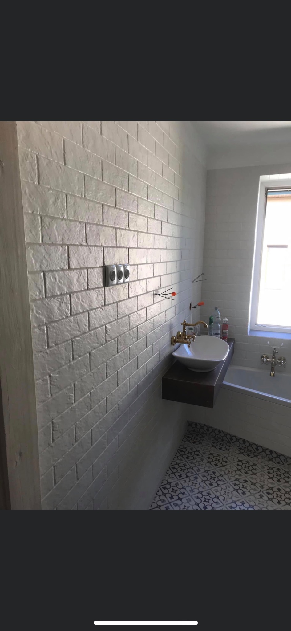 Biała łazienka z płytkami imitującymi cegłę, wisząca umywalka z złotą armaturą na tle wanny i okna, podłoga w czarno-białe wzory.