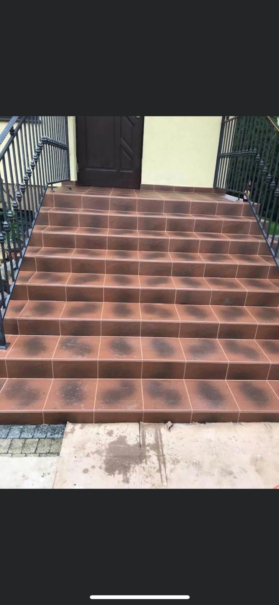 Schody wejściowe wyłożone brązowymi płytkami ceramicznymi z ciemniejszymi akcentami, czarna metalowa balustrada po lewej stronie, ciemne drzwi wejściowe w tle.