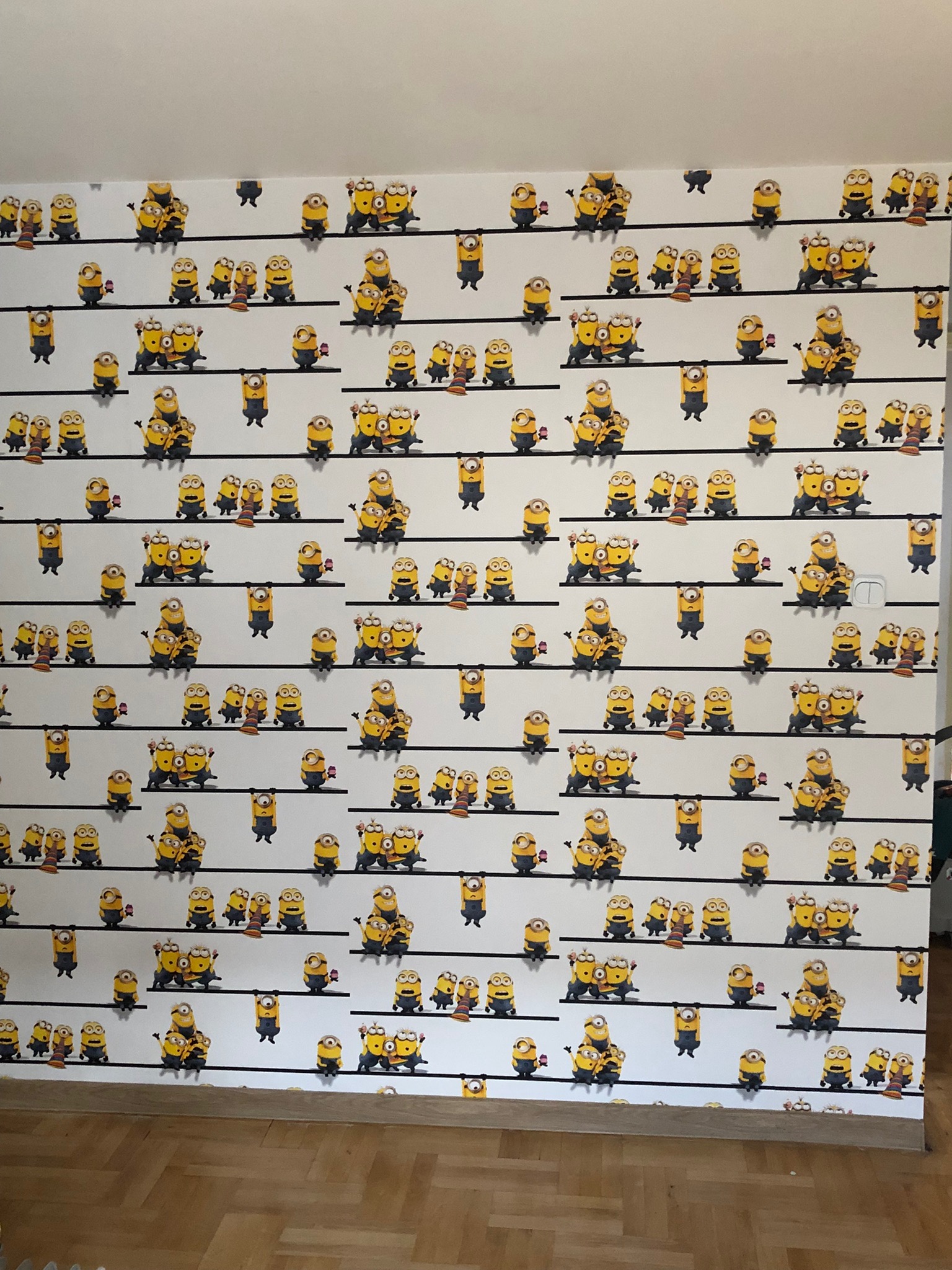 Ściana pokryta tapetą z motywem Minionków na czarnych liniach, widoczny włącznik światła i drewniana podłoga.