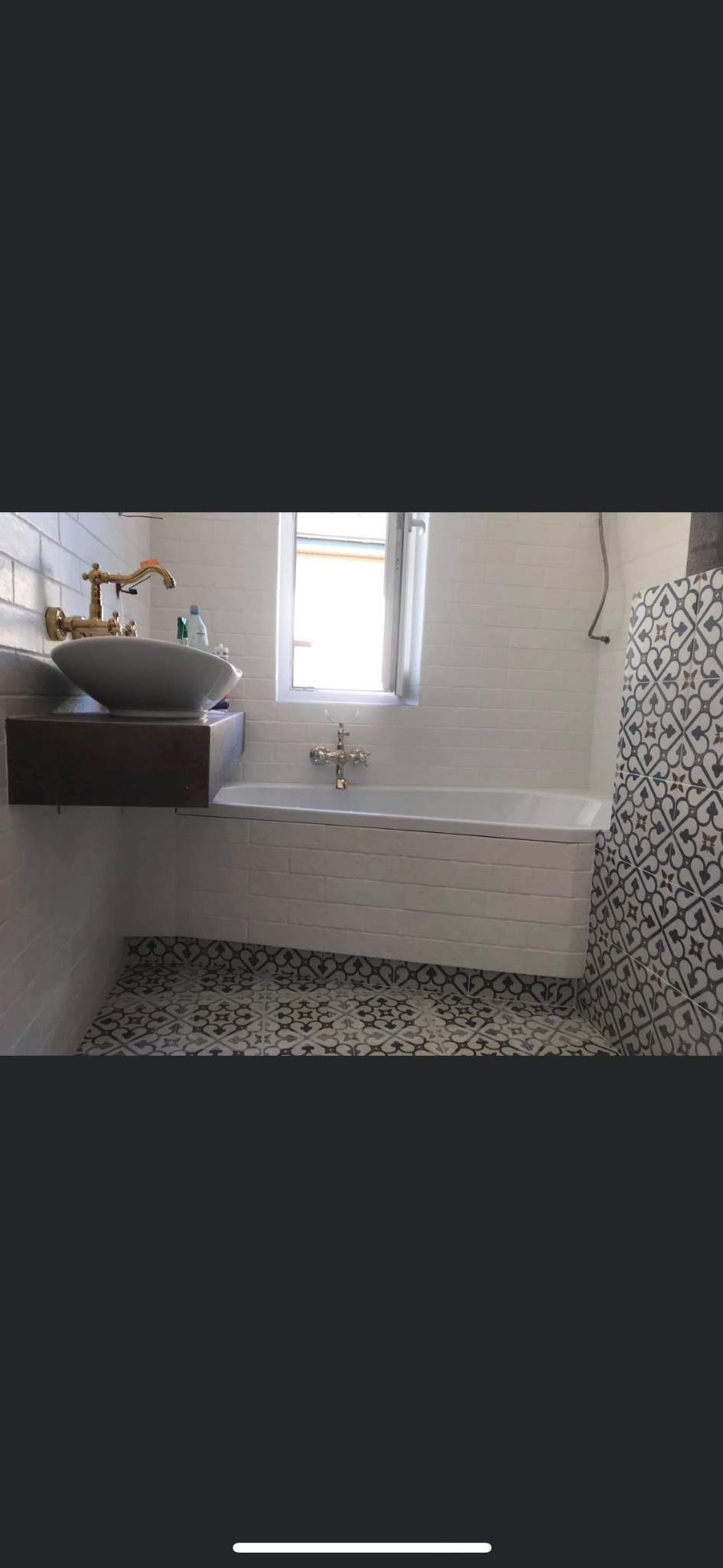 Nowoczesna łazienka z białą cegłą na ścianach, wolnostojącą umywalką na drewnianym blacie, baterią w kolorze złota, białą wanną i wzorzystą, czarno-białą podłogą z płytek.