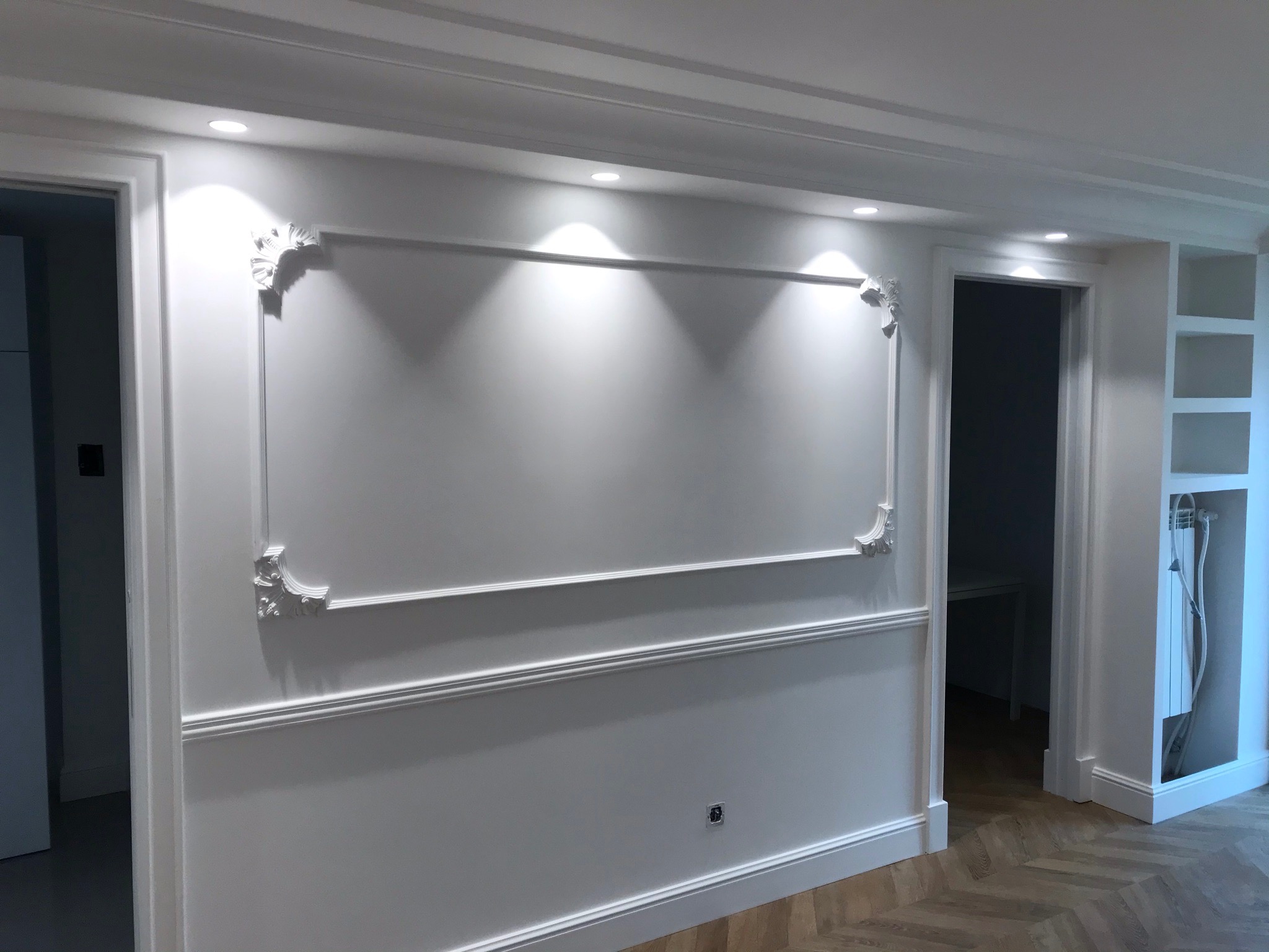 Biała ściana z dekoracyjną sztukaterią w formie ramy i ornamentów narożnych, oświetlona punktowymi lampami sufitowymi; podłoga wyłożona parkietem w jodełkę.