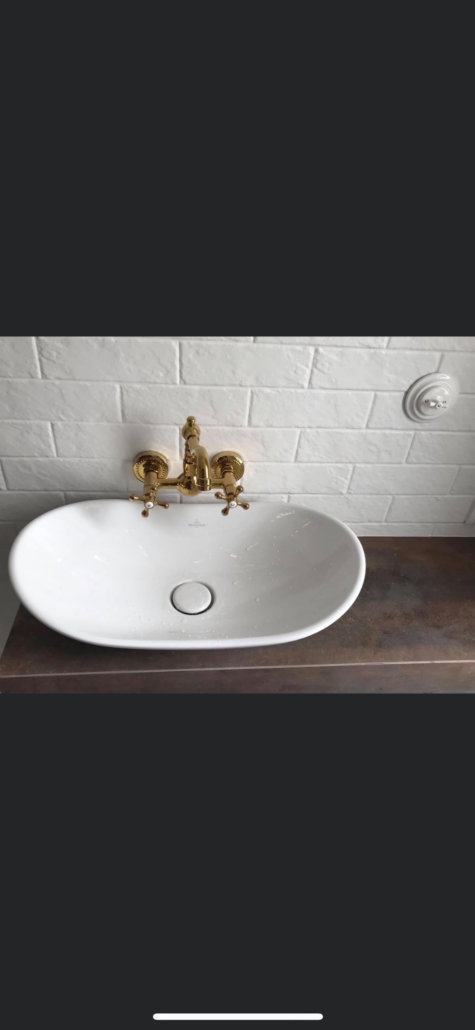 Biała, owalna umywalka Villeroy & Boch z baterią w kolorze złotym na blacie w odcieniach brązu, biała ściana z cegiełek w tle, widoczne krople wody.