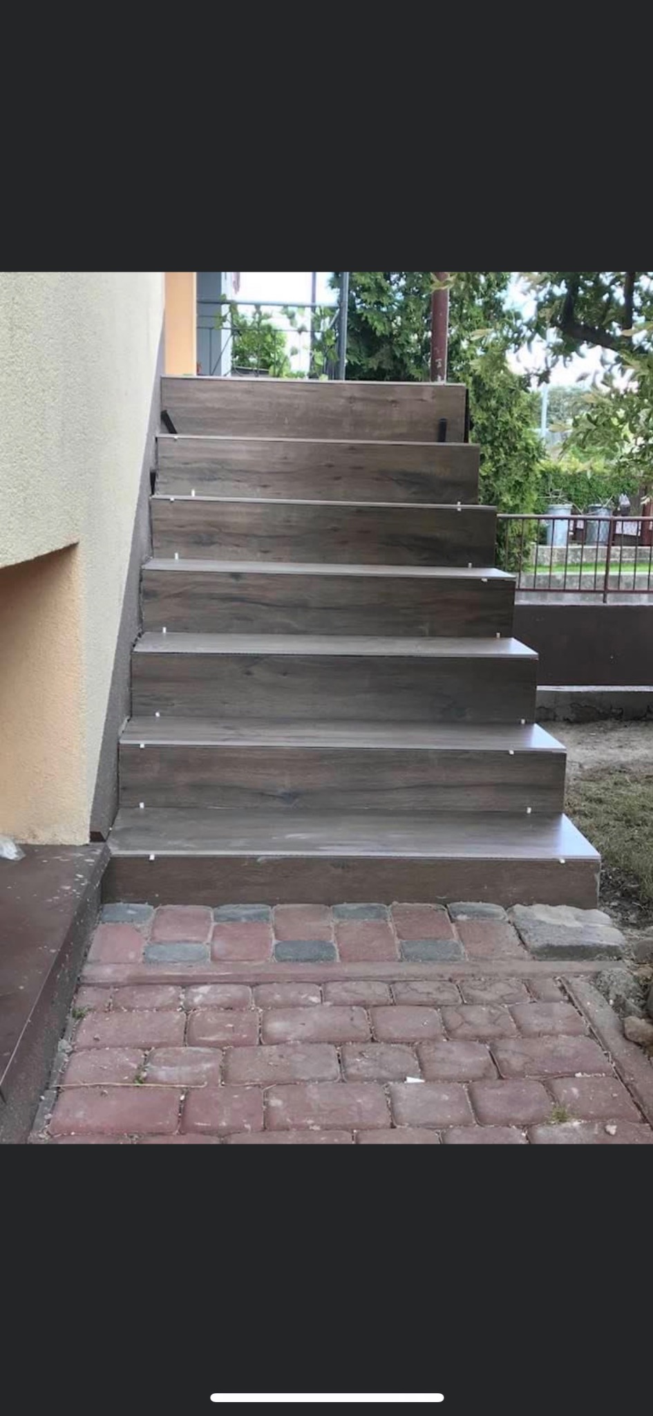 Schody zewnętrzne wyłożone brązowymi płytkami imitującymi drewno, z widocznymi krzyżykami dystansowymi, prowadzące z chodnika z czerwonej kostki.