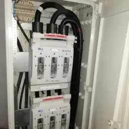 Otwarta skrzynka elektryczna z bezpiecznikami RBK2 i grubymi kablami w peszlach, widoczne złącza i elementy montażowe.