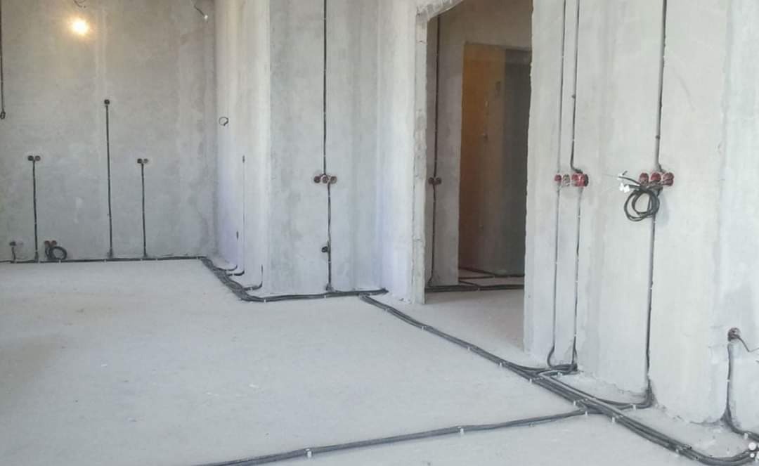 Surowy stan wykończeniowy wnętrza z widoczną instalacją elektryczną w postaci kabli poprowadzonych po ścianach i podłodze, przygotowanych do montażu gniazdek i włączników.