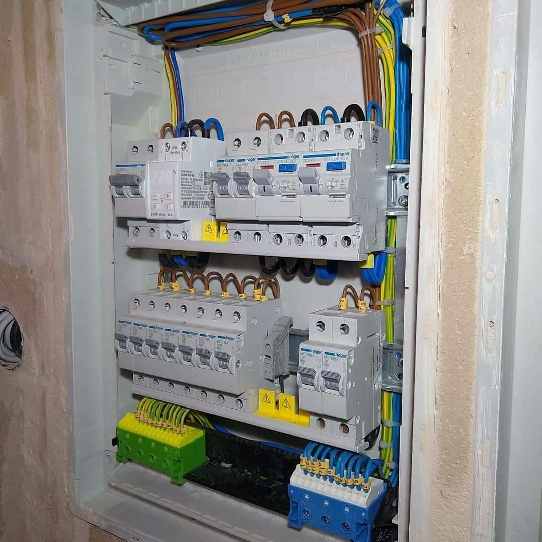 Rozdzielnia elektryczna Hager z zamontowanymi bezpiecznikami i przewodami w kolorach niebieskim, brązowym, żółto-zielonym, widoczne żółte oznaczenia ostrzegawcze.