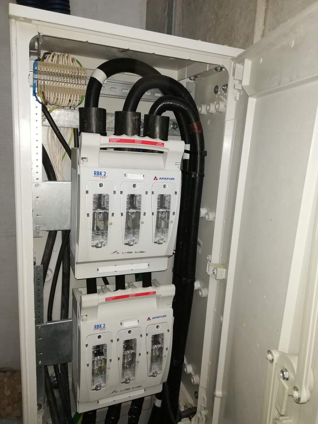 Otwarta skrzynka elektryczna z bezpiecznikami RBK2 i grubymi kablami w peszlach, widoczne złącza i elementy montażowe.