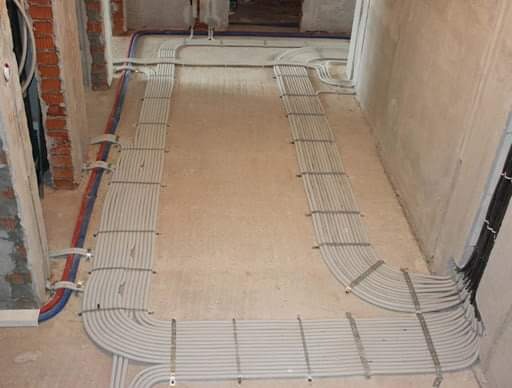 Równolegle ułożone rury instalacyjne, częściowo przymocowane do ściany z surowej cegły, prowadzone wzdłuż podłogi w trakcie remontu.