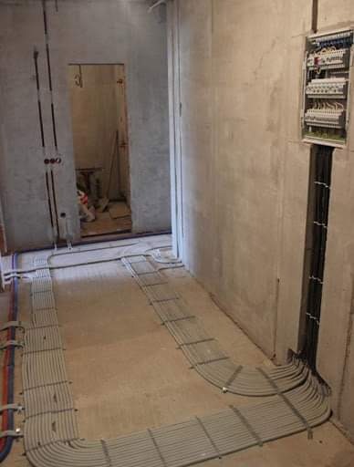 Wnętrze w surowym stanie z rozłożonymi kablami elektrycznymi na podłodze, prowadzącymi do skrzynki rozdzielczej na ścianie. Widoczne pionowe korytka kablowe w ścianie i instalacja podtynkowa.