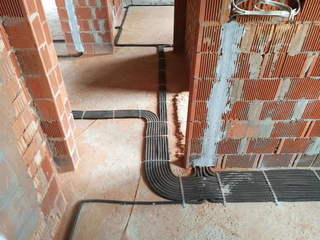 Kable elektryczne w peszlach, prowadzone po podłodze i ścianach w surowym stanie budynku, mocowane metalowymi uchwytami do cegieł.