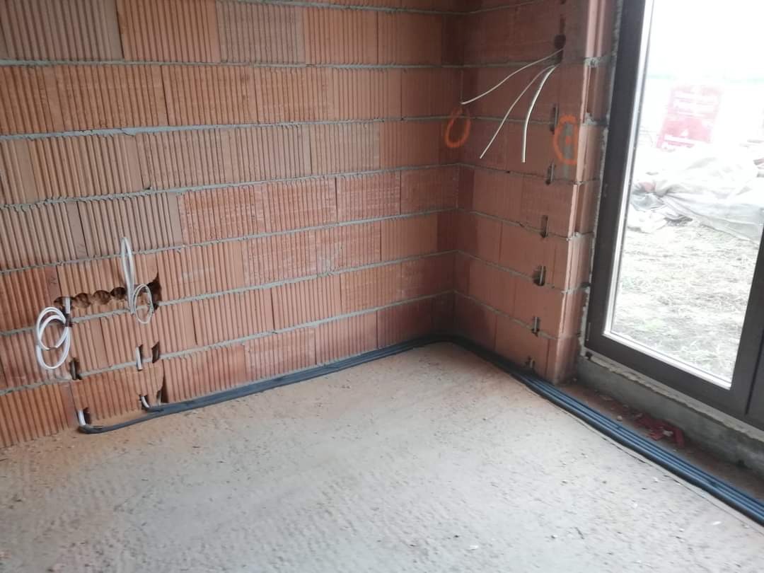 Wnętrze w trakcie budowy z widocznymi przewodami elektrycznymi wystającymi ze ściany z cegły, czarne rurki ochronne prowadzone wzdłuż podłogi, okno w tle.