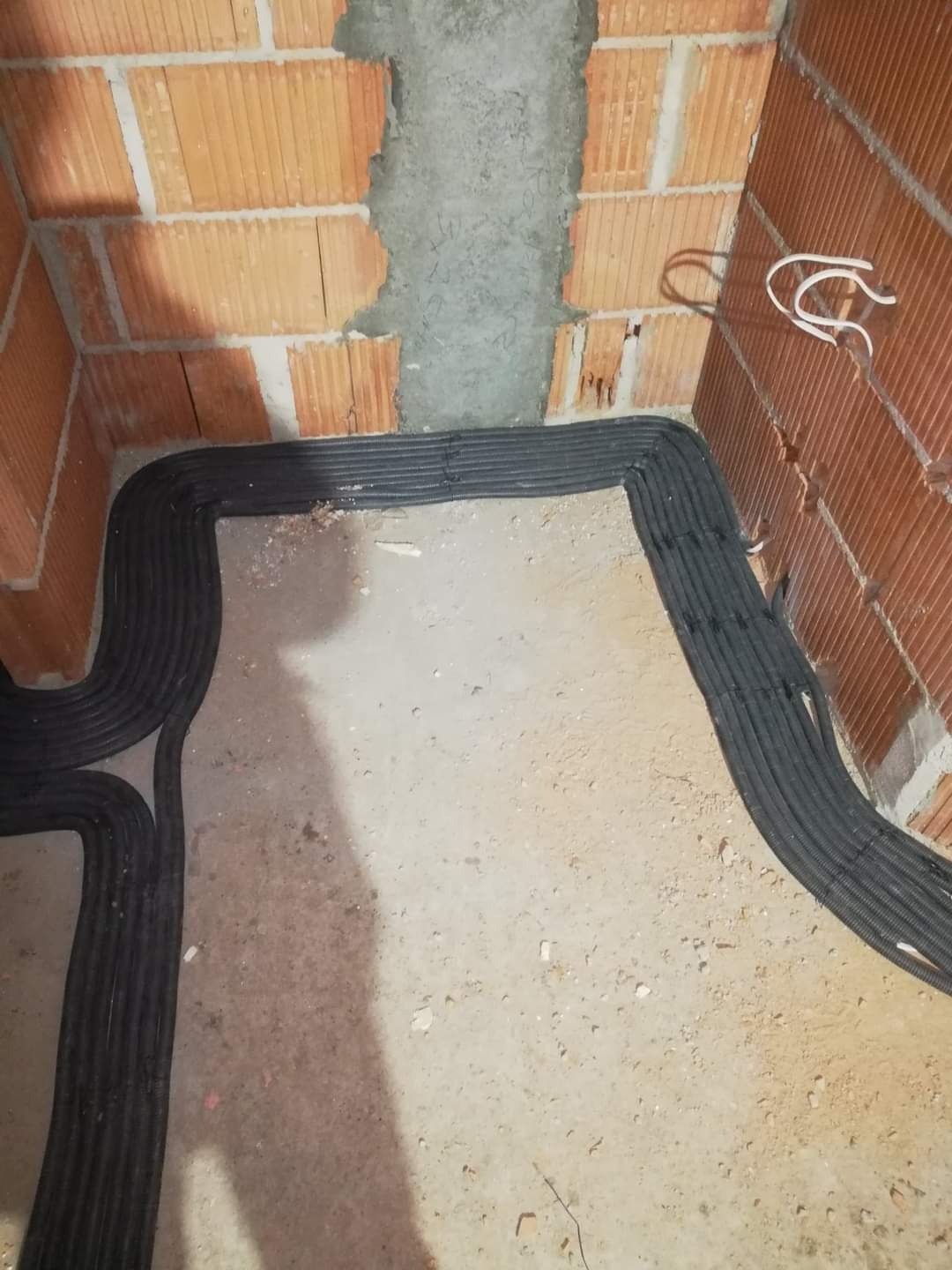 Wnętrze w trakcie prac instalacyjnych: widoczne kable elektryczne w czarnych peszlach ułożone wzdłuż ścian z cegły, częściowo otynkowanych.