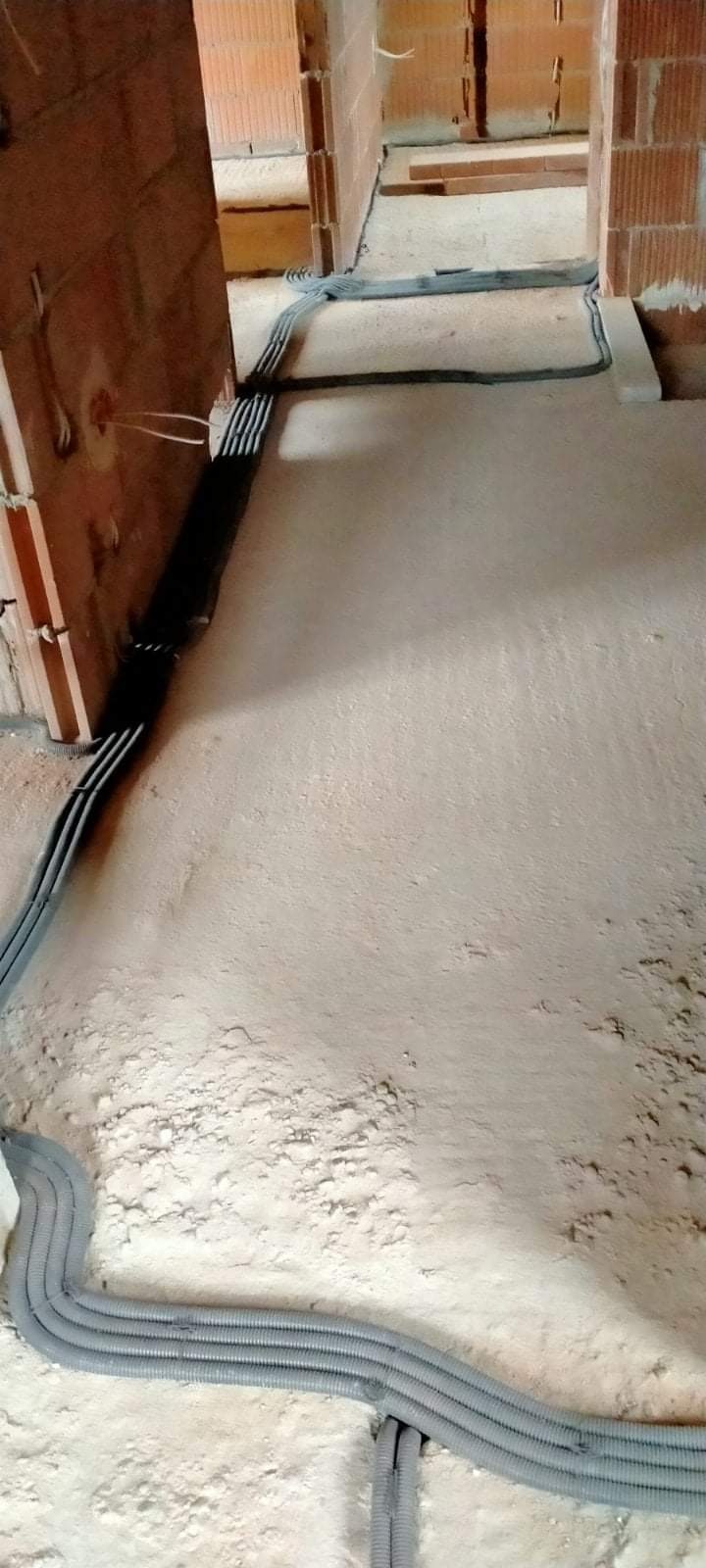 Szare rury karbowane prowadzone po podłodze w surowym stanie, wzdłuż ścian z cegły, instalacja w trakcie budowy.