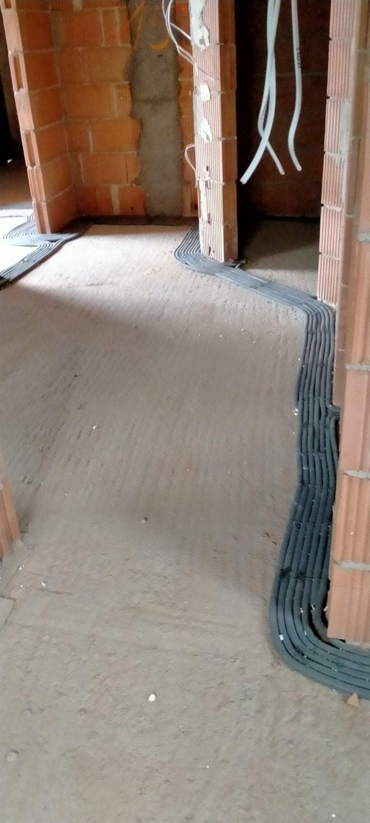 Szare rury ochronne ułożone w bruzdach w podłodze z surowego betonu, prowadzące wzdłuż ceglanych ścian w surowym stanie.