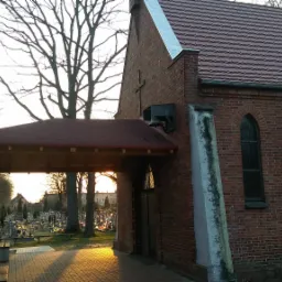 Ceglany budynek z czerwonym dachem i zadaszonym wejściem, widok z zewnątrz, z cmentarzem w tle, oświetlony zachodzącym słońcem.