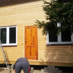 Drewniany dom w trakcie budowy, widoczna elewacja z desek, okna z białymi ramami, drewniane drzwi, pracownicy w ubraniach roboczych i rusztowanie.