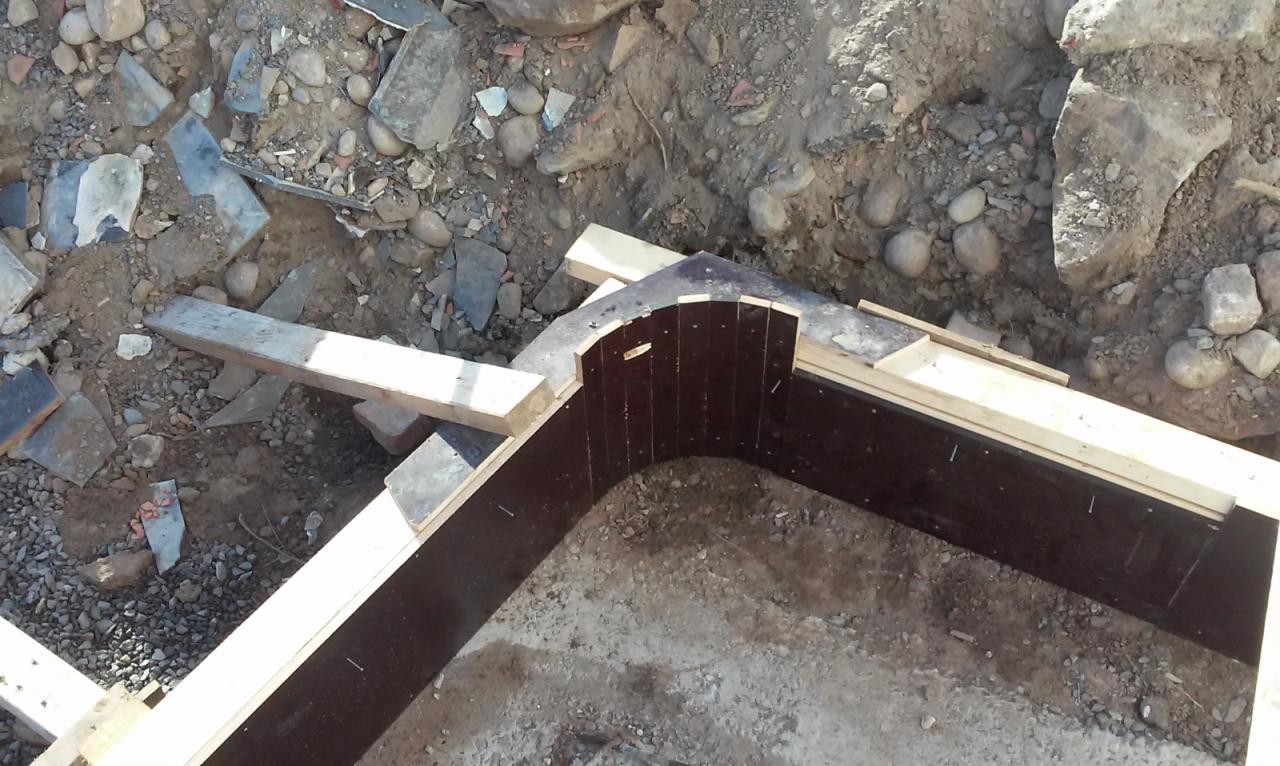 Szczegółowy widok szalunku z desek i płyty na fundamencie o nieregularnym kształcie, przygotowanego do zalania betonem, widoczne kamienie i gruz w tle.