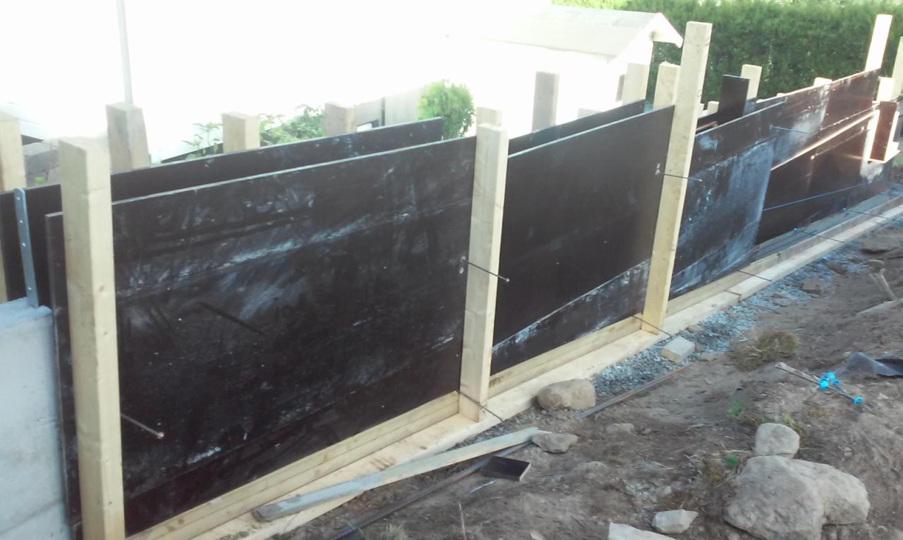 Szalunek z czarną folią i drewnianymi stemplami, przygotowany do wylania fundamentów, widoczne zbrojenia i kamienie w tle.