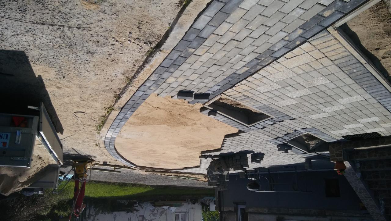 Układanie szarej kostki brukowej na podsypce piaskowej, widoczne krawężniki w ciemniejszym odcieniu i trawnik w tle.