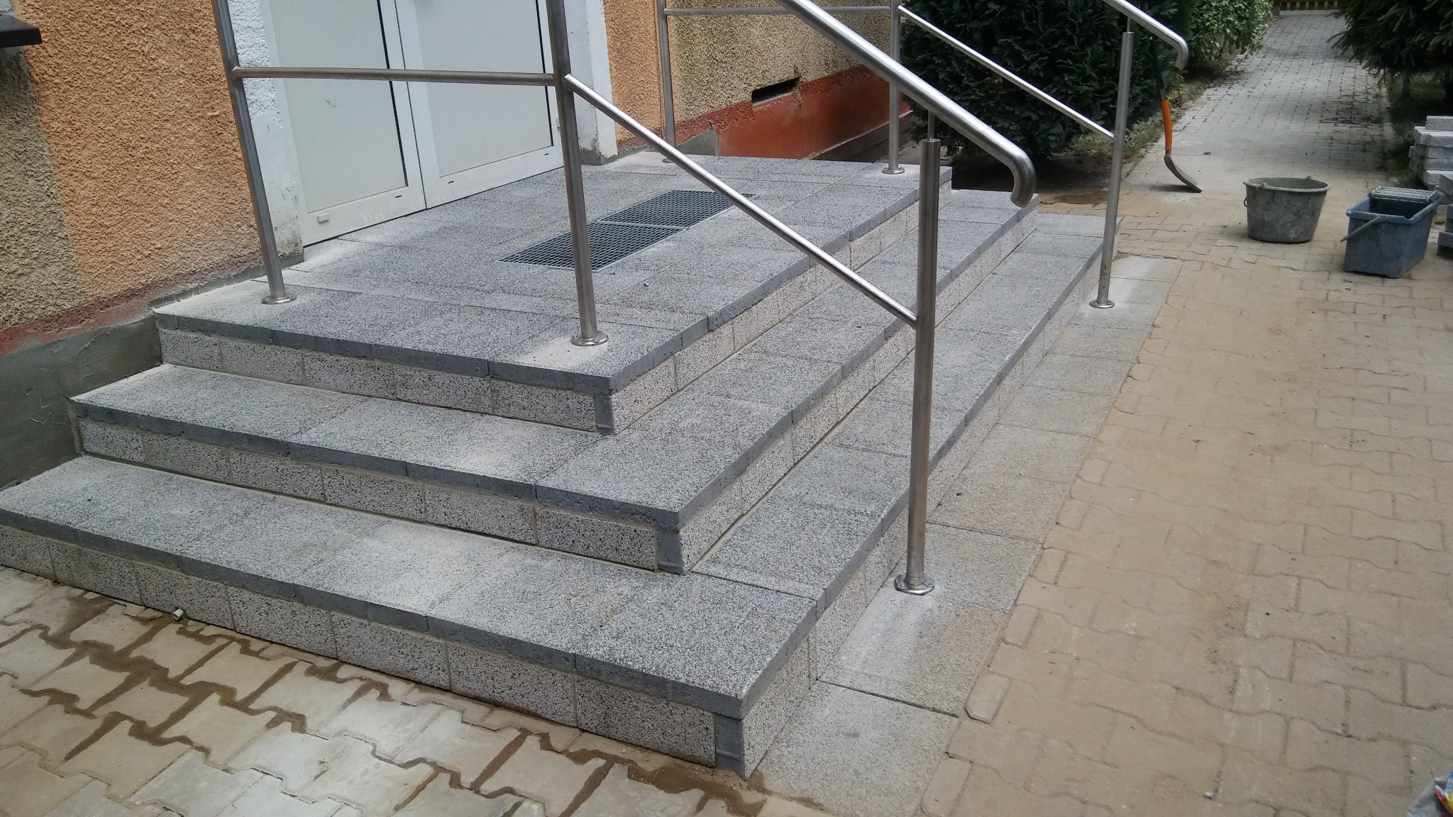 Schody wejściowe wyłożone szarą kostką granitową z metalową balustradą, widoczne dwa wiadra i łopata w tle.
