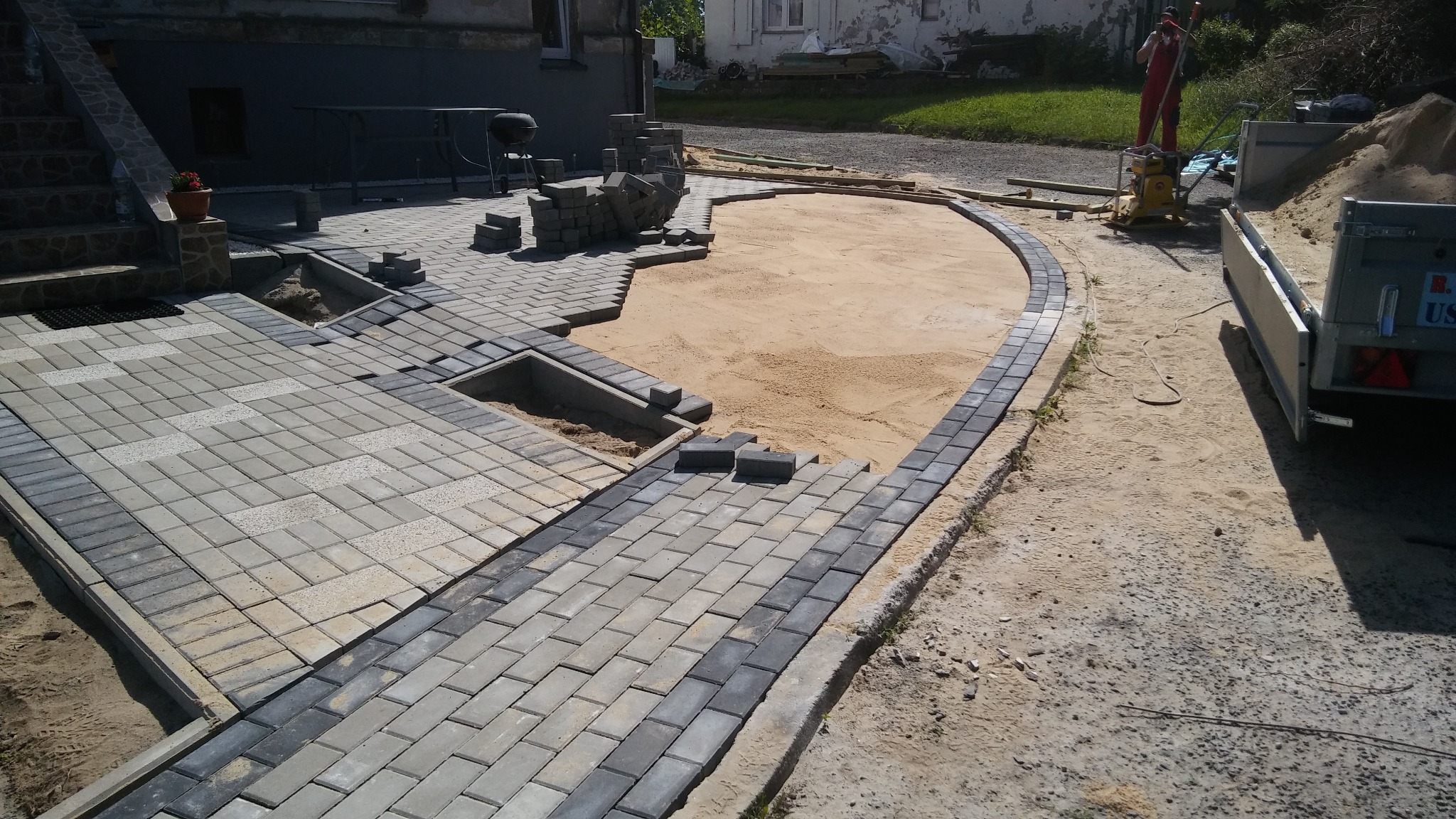 Układanie szaro-białej kostki brukowej na podsypce piaskowej, widoczny fragment schodów, trawnik i pracownik ubijający piasek zagęszczarką w tle.