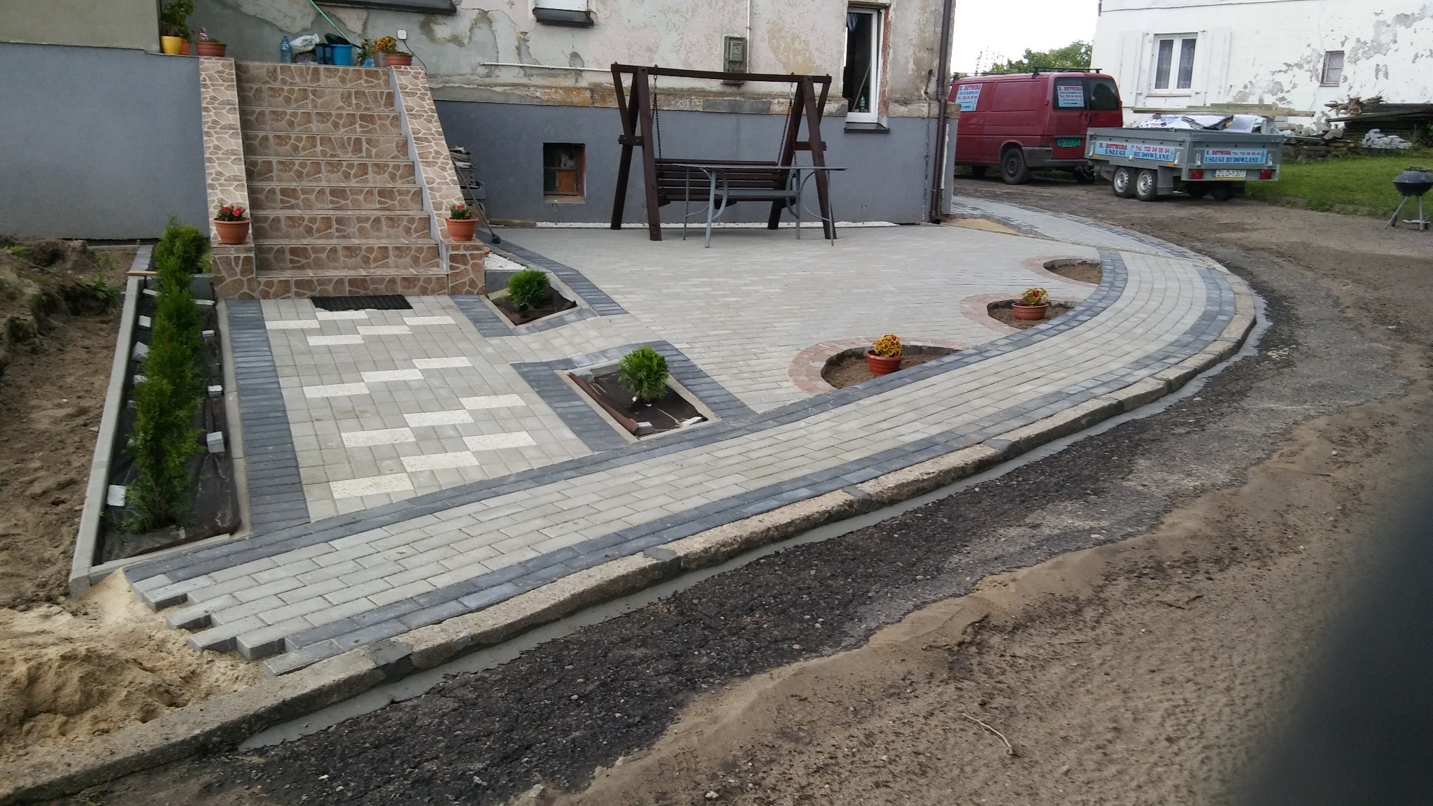 Ułożona kostka brukowa w odcieniach szarości i bieli tworząca ścieżkę i taras z elementami dekoracyjnymi w postaci klombów z roślinami i schodów wejściowych do budynku, w tle huśtawka i samochód...
