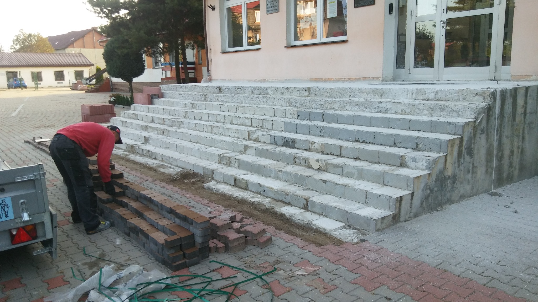 Pracownik w czerwonej bluzie układa ceglaną kostkę klinkierową na schodach wejściowych do budynku, widoczny fragment przyczepki i narzędzia.