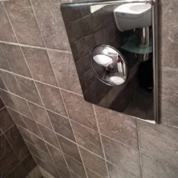 Błyszczący, chromowany panel spłuczki toalety z dwoma przyciskami, zamontowany na ścianie z brązowych, kwadratowych płytek; w odbiciu panelu widoczne umywalka i fragment wnętrza łazienki.