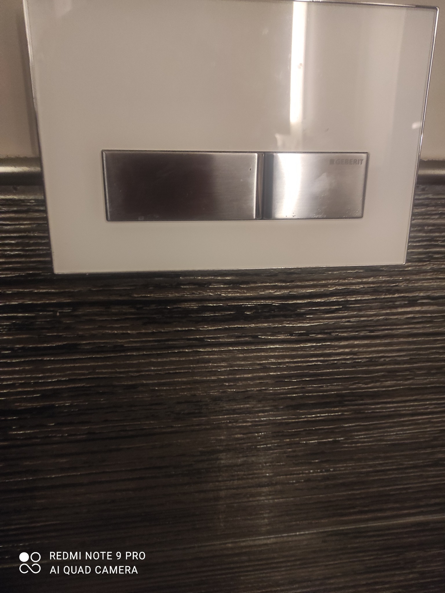 Przycisk spłuczki Geberit na białej płytce, zamontowany na brązowej ścianie o teksturze drewna.