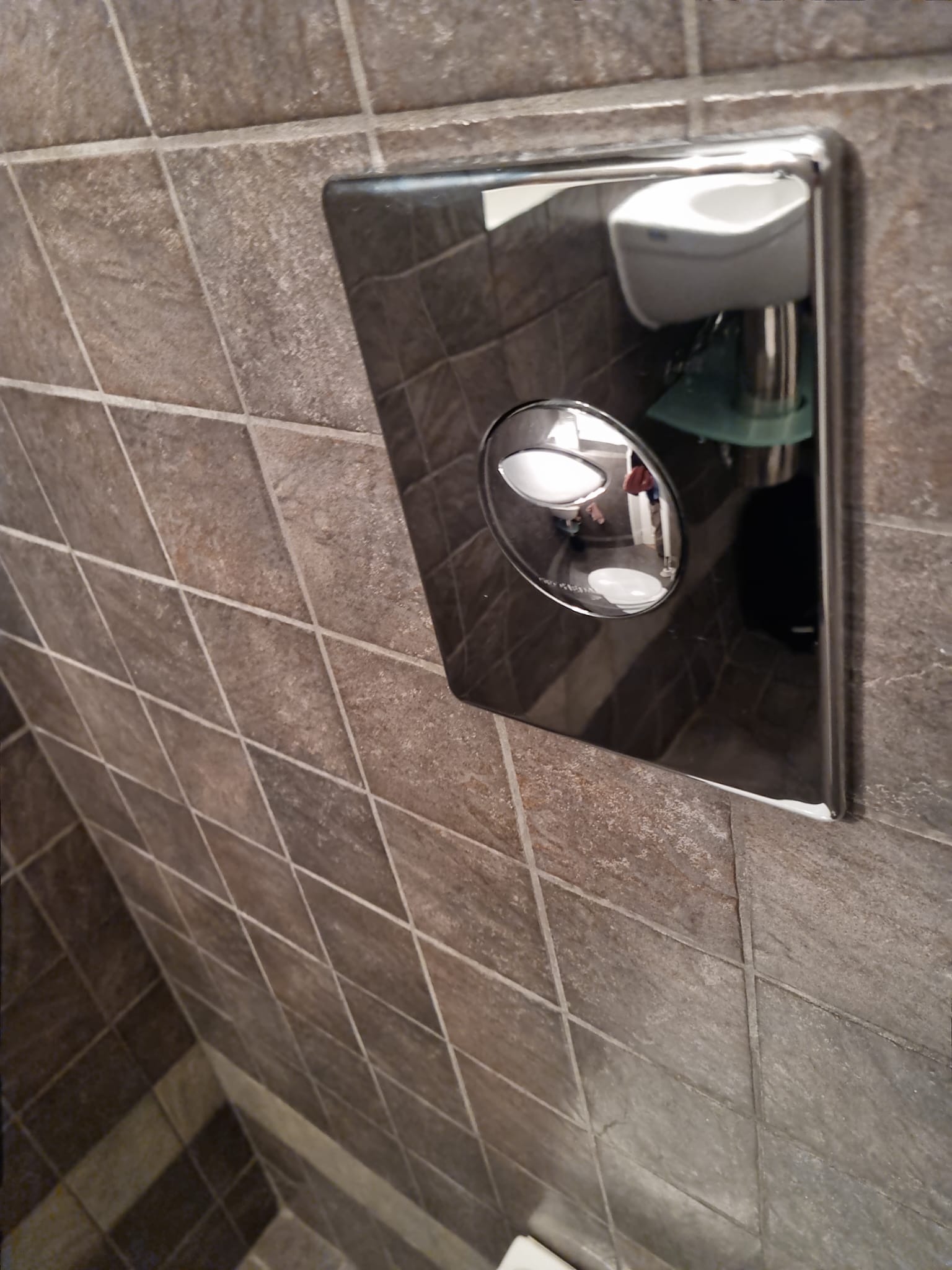 Błyszczący, chromowany panel spłuczki toalety z dwoma przyciskami, zamontowany na ścianie z brązowych, kwadratowych płytek; w odbiciu panelu widoczne umywalka i fragment wnętrza łazienki.