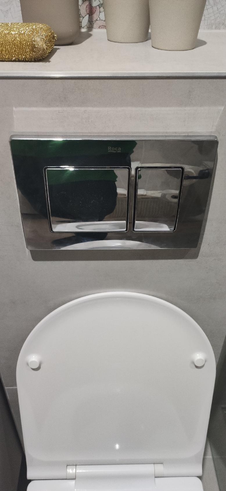 Zamontowana spłuczka Roca z dwoma przyciskami na szarej płytce ceramicznej nad białą deską sedesową, widoczne odbicie otoczenia w chromowanej powierzchni.