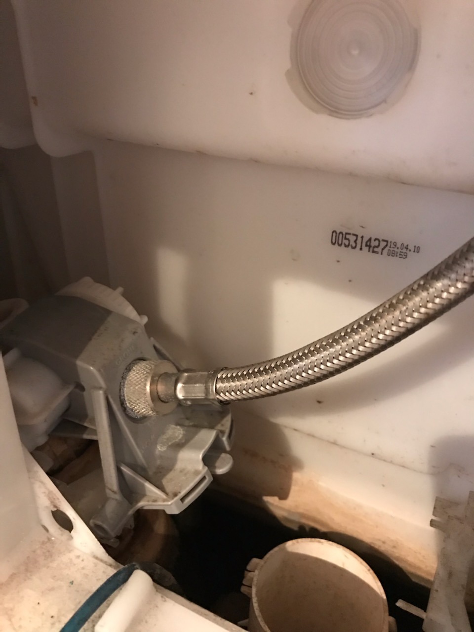 Wnętrze spłuczki toalety z widocznym mechanizmem spłukującym, przyłączem wężykowym w oplocie metalowym i datą produkcji na obudowie.