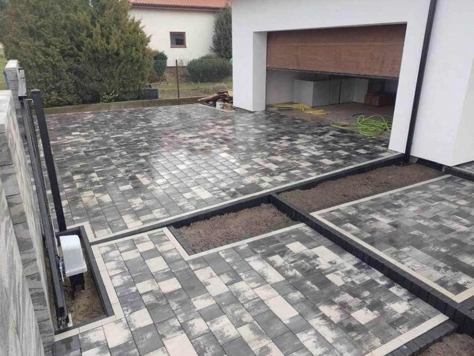 Ułożona, dwukolorowa kostka brukowa na podjeździe z wyznaczonymi miejscami na zieleń, widoczny otwarty garaż i częściowo brama wjazdowa.