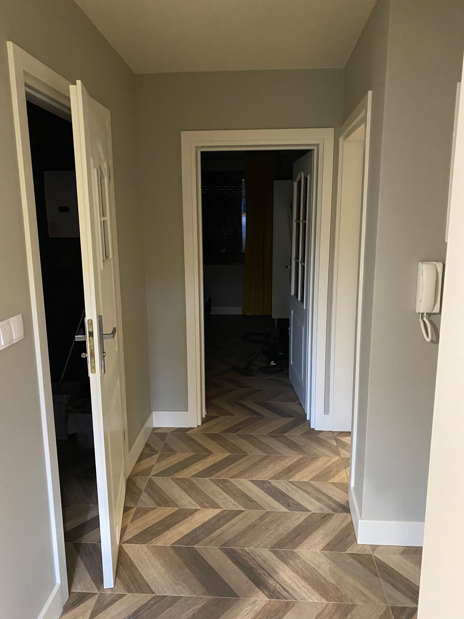 Wnętrze mieszkania z ułożoną podłogą w jodełkę, widok na otwarte drzwi do kolejnych pomieszczeń, ściany w odcieniach szarości.