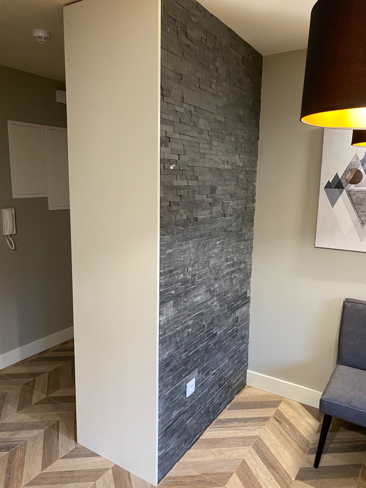 Fragment wnętrza z dekoracyjną ścianą z szarego kamienia łupkowego, widoczny fragment białej zabudowy, parkiet w jodełkę oraz szare krzesło i lampa z abażurem.