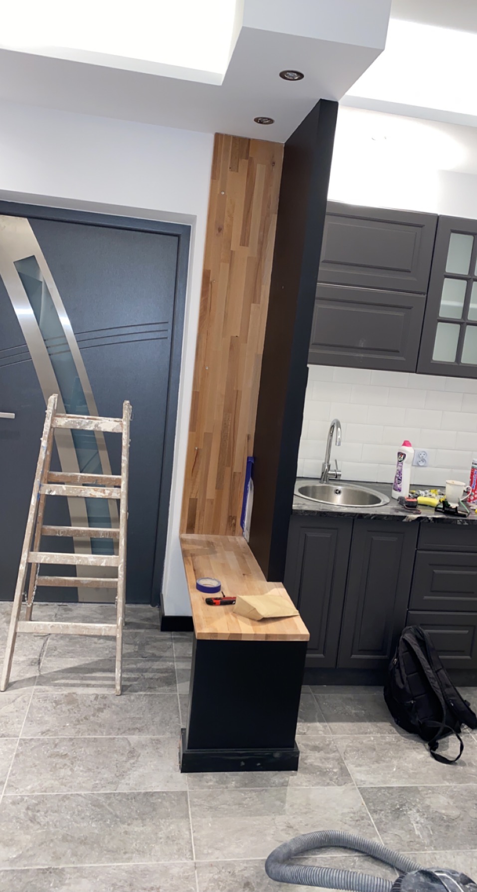 Wnętrze w trakcie prac wykończeniowych: drabina oparta o ścianę, fragment drewnianej boazerii, zabudowa kuchenna w tle, widoczny fragment odkurzacza na podłodze.