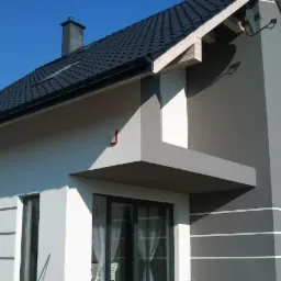 Nowoczesny dom jednorodzinny z elewacją w odcieniach szarości i bieli, wykończony czarną dachówką i elementami dekoracyjnymi w postaci poziomych pasów. Widoczny fragment ogrodu i błękitne niebo.