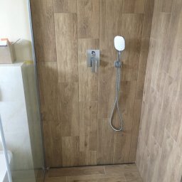 Przemysław Gołębiewski - Kabina prysznicowa wykończona płytkami imitującymi drewno, z zamontowaną baterią i słuchawką prysznicową na ścianie, widoczny fragment brodzika i szklana ściana kabiny.