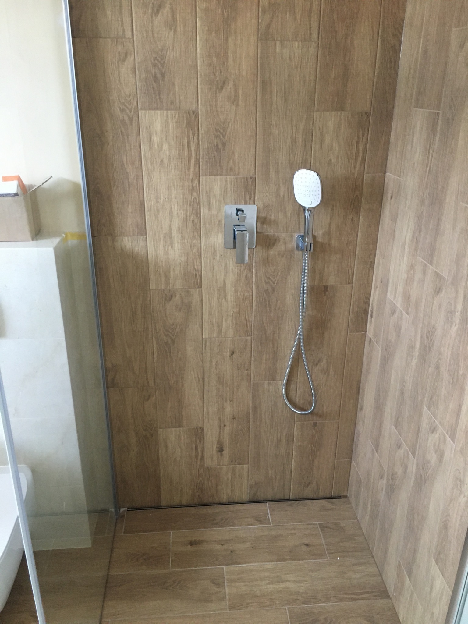 Kabina prysznicowa wykończona płytkami imitującymi drewno, z zamontowaną baterią i słuchawką prysznicową na ścianie, widoczny fragment brodzika i szklana ściana kabiny.