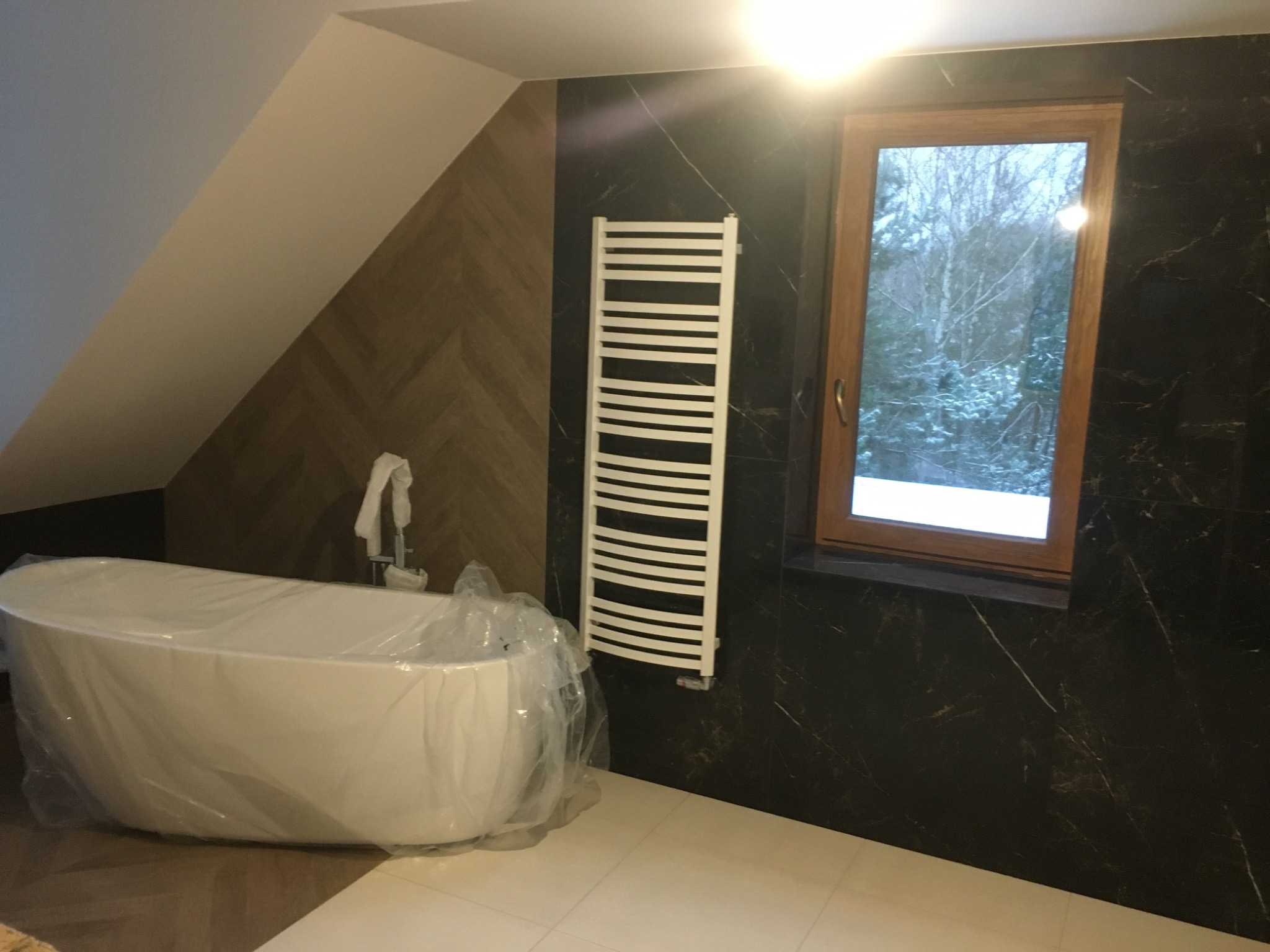 Nowoczesna łazienka w trakcie wykończenia: wolnostojąca wanna owinięta folią ochronną, grzejnik drabinkowy na czarnej ścianie imitującej marmur, okno z widokiem na zimowy krajobraz.