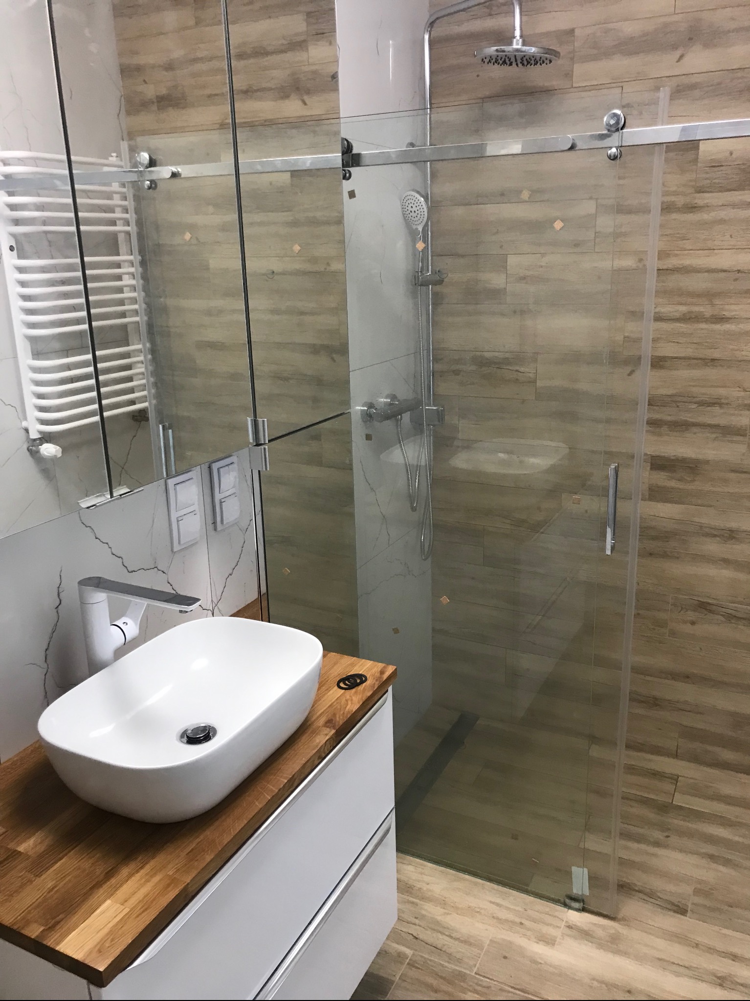 Nowoczesna łazienka z kabiną prysznicową ze szklanymi przesuwnymi drzwiami, umywalką nablatową na drewnianym blacie i grzejnikiem drabinkowym.