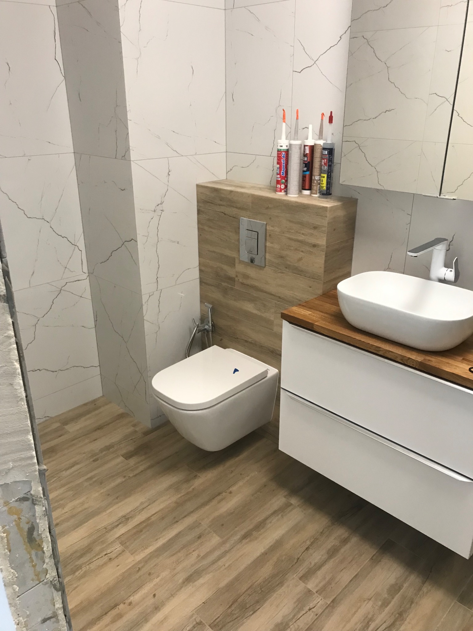 Nowoczesna łazienka z białymi płytkami imitującymi marmur, drewnopodobną podłogą, podwieszaną toaletą i umywalką nablatową na blacie z drewna. Widoczne tuby z silikonem na zabudowie stelaża.
