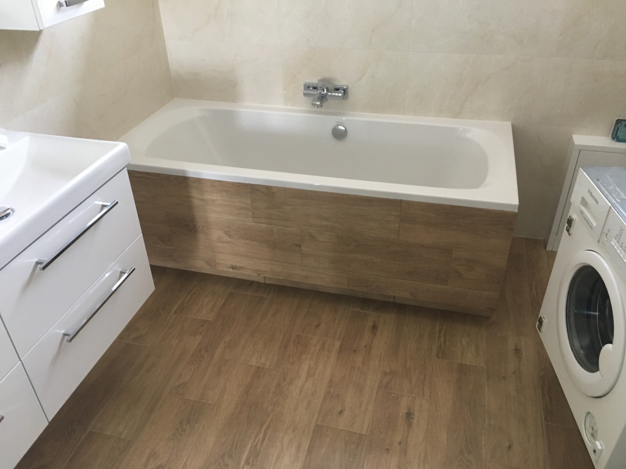 Nowoczesna łazienka z białą wanną obudowaną panelami imitującymi drewno, białą umywalką z szafką i pralką. Podłoga wyłożona panelami podłogowymi w odcieniach brązu.