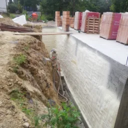 Pracownik w kombinezonie ochronnym aplikuje białą pianę izolacyjną na ścianę fundamentową wykopu, z widocznymi przewodami i drabiną, w tle palety z cegłami Wienerberger Porotherm.