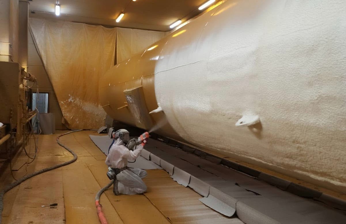 Pracownik w kombinezonie ochronnym natryskuje pianę izolacyjną na duży zbiornik wewnątrz budynku. Widoczne węże i osłony podłogowe.