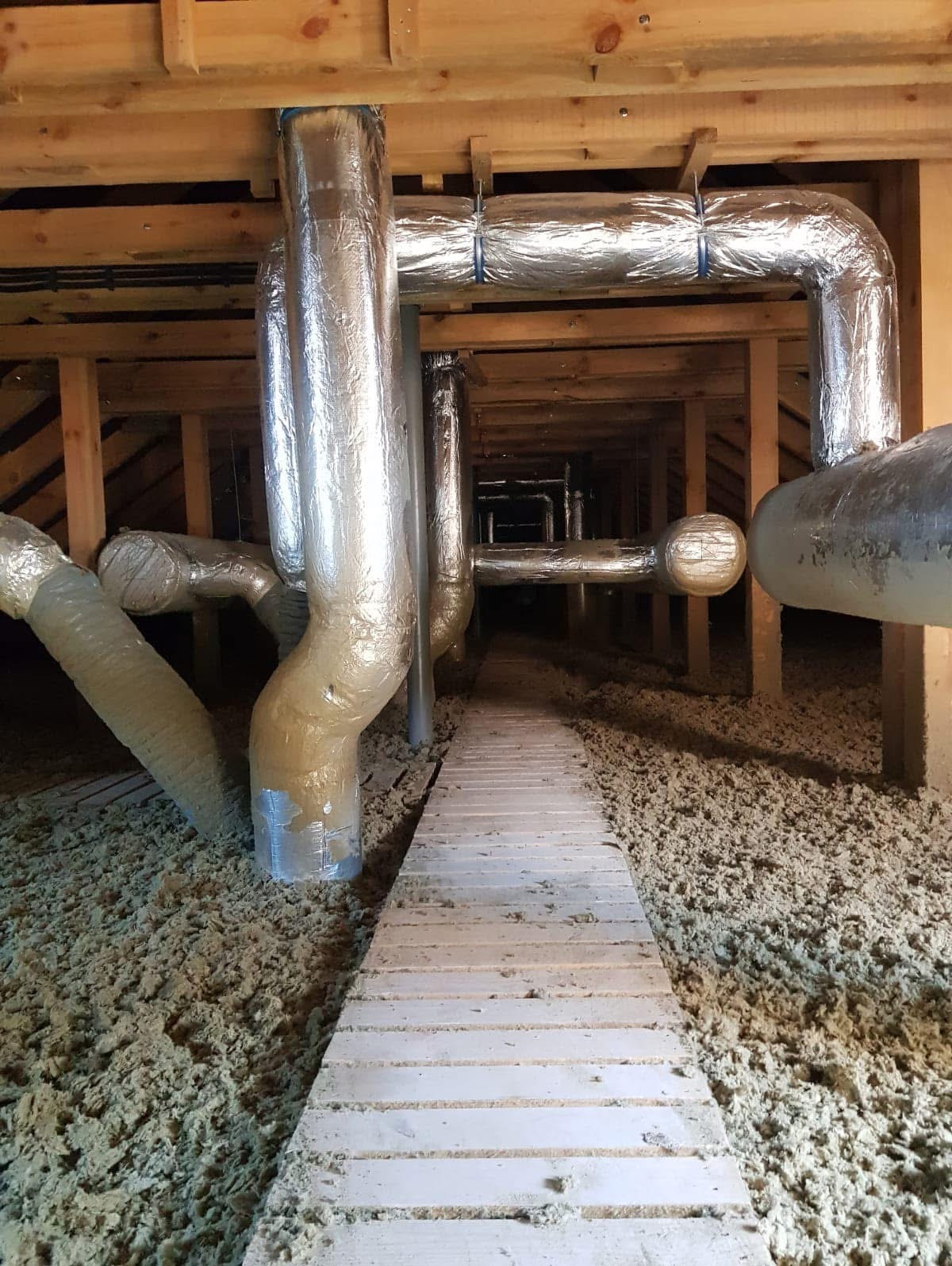 Izolacja poddasza wełną mineralną, widoczne rury wentylacyjne w srebrnej folii. Drewniana konstrukcja dachu i wąska ścieżka z desek.