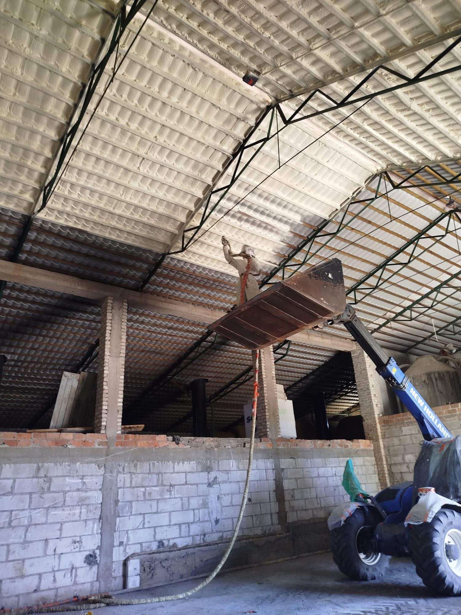 Pracownik w kombinezonie nanosi izolację piankową na metalową konstrukcję dachu wewnątrz budynku. Widoczny podnośnik i fragment ściany z cegieł.